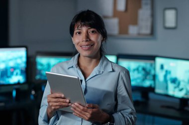 Genç başarılı gözetleme çalışanı tablet kullanıyor ve bilgisayar monitörlerinin karşısında dururken kameraya bakıyor.