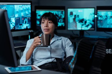 Bir fincan çay ya da kahve içen genç, ciddi bir kadın iş yerinde bilgisayar monitörünün önünde oturuyor ve ekranda CCTV videosu izliyor.