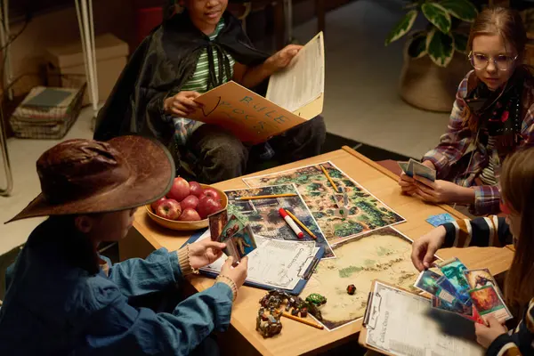 stock image High angle view of several youthful intercultural kids in costumes sitting by table during tabletop game while boy putting one of his cards on board