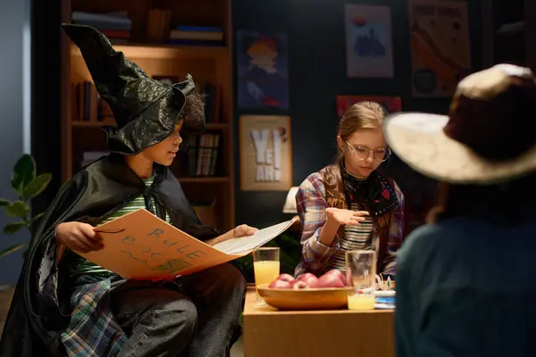stock image Cute African American boy in witch hat and mantle sitting next to youthful girl during board game and explaining rules from big book