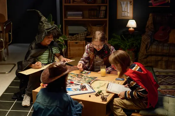 stock image Four intercultural youthful children in costumes of board game characters sitting by table and bending over pictures with figurines