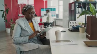 Orta boy Afro-Amerikan reklam müdürü, kreatif ajans masasında oturan etnik türban, sunum sayfasındaki verilere bakarak, dijital tablet üzerinde yazarken.