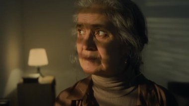 Yas tutan yaşlı beyaz kadının yakın plan portresi. Şafak vakti karanlık odada üzgün görünüyor ve kameraya dönüyor.