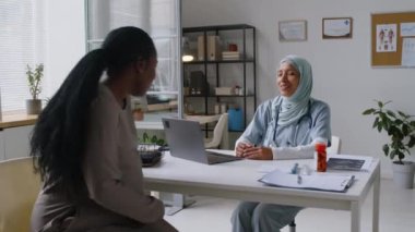 Sağlık kontrolü için GP ofisine giren Afrikalı hamile bir kadının orta boy fotoğrafı. İslami başörtüsü takmış kadın doktor onu selamlıyor, refah ve şikayetleri soruyor.