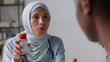 İslami tesettürde gülümseyen Müslüman doktorun GP kliniğindeki kimliği belirsiz erkek veya kadın hastaya reçeteli ilaç verirken orta boy yakın plan çekimleri, potansiyel yan etkiler hakkında uyarıda bulundu.