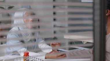 Klinikte oturan, hamile siyahi bir kadını teselli eden, doğum öncesi kontroller sırasında elini tutup okşayan sempatik, genç Müslüman kadın doktorun pencere arkasından çekimleri.