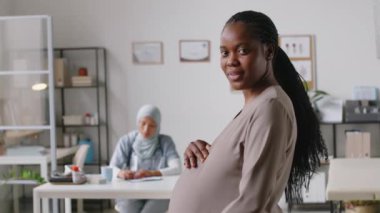 Doğum öncesi muayenesi için klinikte bekleyen mutlu, gülümseyen Afrikalı Amerikalı genç bir kadının orta boy portresi, arka planda evrakları dolduran Müslüman doktor.