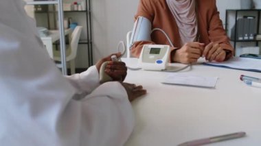 Tanınamayan Afrikalı Amerikalı doktorun klinikte Müslüman kadın hastayı tedavi ederken, ampul pompalarken, kan basıncını ölçerken, kadın soru sorarken orta boy fotoğrafı.