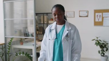 Üniformalı, beyaz önlüklü, eldivenli, göğüs göğüste duran ve kameraya özgüvenle bakan Afrikalı Amerikalı kadın pratisyenin orta boy portresi.