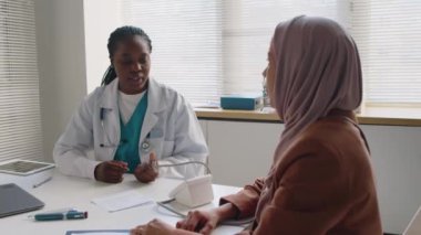 Siyahi kadın hekimin klinikte masasında oturup, başörtüsü takan Müslüman hastaya kan basıncını ölçerken aynı zamanda semptomlar, şikayetler ve tıbbi geçmişle ilgili sorular sorduğu orta boy bir fotoğraf.