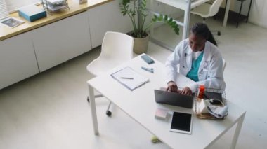 Afrika kökenli Amerikalı bir kadın doktorun klinikte oturduğu, dizüstü bilgisayarla çalıştığı, tesettürlü Müslüman bir hastanın içeri girip, oturduğu ve migrenden şikayet ettiği tam açılı bir fotoğraf.