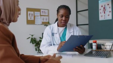 Siyahi kadın klinisyenlerin evrak kutularını işaretlerken, imzalarken, sonra da başörtüsü takan Müslüman hastalara ameliyat ya da prosedür için evrak hazırlarken orta boy bir fotoğraf.