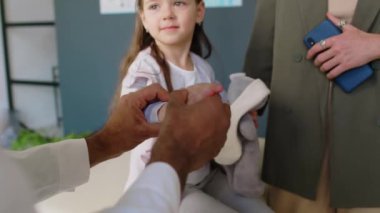 Yetişkin erkek travmatoloğun ellerinin yakın plan çekimi. Burkulma ya da yaralanmadan sonra 6 yaşındaki kızları muayene ediyor. Parmaklarını ve bileklerini oynatıyor, GP ameliyatındaki randevusu sırasında ağrıyı soruyor.