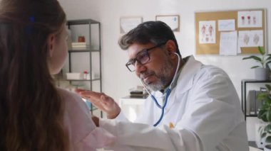 Hintli erkek çocuk doktorunun orta ölçekli görüntüleri kanepede oturan beyaz kızı muayene ediyor, steteskopla göğsünü dinliyor, nefes almak ve vermek istiyor, gözleri kontrol ediyor,
