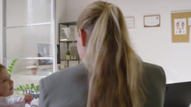 Orta boy Hintli erkek aile doktorunun klinikte masasında oturup, beyaz anne ve kızına danışarak göğüs röntgenini kız tablete gösterip, şakacı bir şekilde anlamını açıkladığı bir fotoğraf.