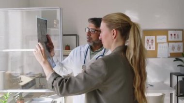 Yetişkin Asyalı erkek nörologların orta boy fotoğrafları ve klinikteki pencerenin yanında duran, ışığa karşı filmlerdeki beyin MR görüntülerine bakıp anormallikleri tartışan, sıkıntılı beyaz kadın hastalar.
