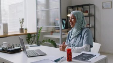 Müslüman bir kadının tesettürlü ve doktor üniformalı bir şekilde klinikte otururken, dizüstü bilgisayardaki düğmeye basarken, hastayla internetten canlı bir video görüşmesi yaparken, gülümserken.