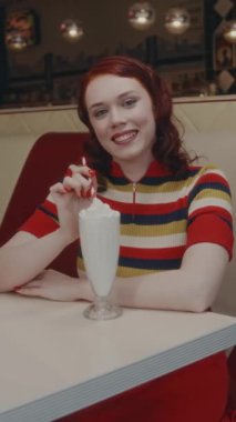 Kırmızı saçlı, çizgili polo tişörtlü çekici beyaz kadının dikey portresi. Klasik lokantada masada milkshake ile poz veriyor ve kameraya bakıyor.