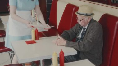 1970 'lerin tarzında otantik bir lokantada, üniformalı, tanınmayan bir garson menüsünü saman şapkalı, beyaz bir beyefendiye getirirken çekilmiş bir fotoğraf.