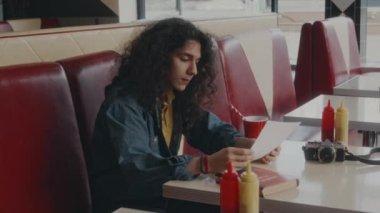 Z jenerasyonundaki kıvırcık saçlı beyaz adamın menüde yemek seçerken soda içerken otantik bir kafede otururken çekilmiş bir fotoğrafı.