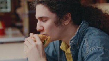 Kot ceketli, uzun saçlı, geçmişe dönük lokantada hamburger yiyen ve hayatın tadını çıkaran mutlu beyaz adamın yakın çekimi.