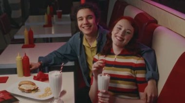 1980 'lerdeki Amerikan kafesinde milkshake ve abur cuburla oturan beyaz bir çiftin orta boy portresi ve gülümseyerek kameraya bakmaları.