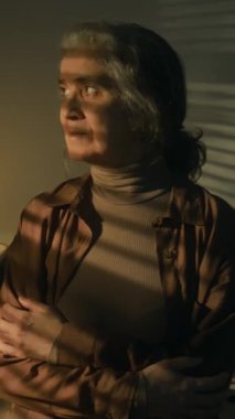 Ciddi bir beyaz kadının dikey portresi. Karanlık odada kollarını kavuşturup kameraya dönüyor.