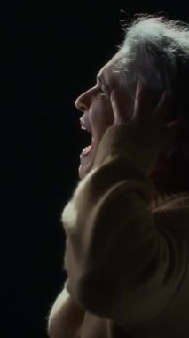 Süveter giymiş yaşlı beyaz bir kadının dikey portresi. Siyah arka plan karşısında el ele tutuşup acı içinde bağırıyor. Umutsuzluk ve umutsuzluk hissediyor.