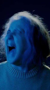 Süveter giymiş yaşlı bir kadının dikey yavaş portresi. Panik içinde gözleri kapalı bağırıyor. Stüdyoda siyah arka planda mavi ışıklı bir kafa sallıyor.