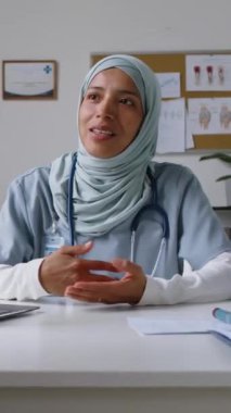 İslami başörtüsü takmış gülümseyen bir kadın doğum uzmanı klinikte oturmuş hamile bir kadına danışıyor ve doğum tarihinden önce bebeğin karnını okşuyor.