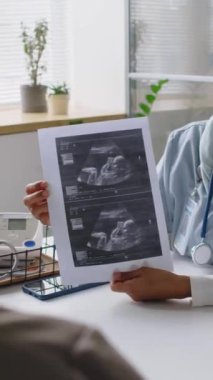 Genç kadın jinekoloğun klinikte ya da doğum hastanesinde oturmuş hamile bir kadına ultrason görüntüsü gösterirken dikey görüntüsü.