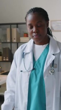 Üniformalı, beyaz önlüklü, eldivenli, kollarını göğsünde birleştirerek kameraya bakıp gülümseyen Afro-Amerikan kadın klinisyenlerin orta boy dikey portresi.