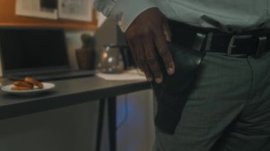 Gri gömlekli, pantolonlu, siyah deri kemerli, elini beline koyan, görev silahıyla tanınmayan Afro-Amerikan polis dedektifini yakından çek.
