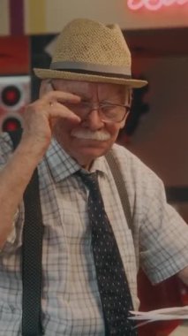 Hasır şapkalı yaşlı beyefendinin dikey yan portresi. Gözlüklerini çıkarıyor ve retro lokantada otururken kameraya bakıyor.