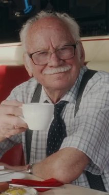 İleri yaşlarda beyaz bir beyefendinin dikey portresi. Gözlük takıyor ve kahve içerken retro tarzı Amerikan lokantasında kahvaltı yapıyor.