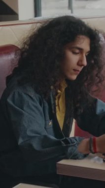 Z Geni 'nin dikey yan görüntüsünde kıvırcık saçlı, menüde yemek seçen ve soda içen otantik bir kafede oturan beyaz adam.