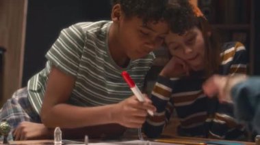 Orta boy Afro-Amerikan çocuğun masaya yaslanıp masa oyunu için konum haritası çizerken çeşitli arkadaşları akşamları onu izlerken.
