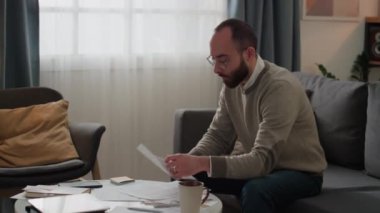 Orta boy Orta Doğulu bir adamın evdeki kanepede oturup ev harcamalarıyla uğraşırken, zarfı açarken, yeni bir fatura çıkarırken, onu incelerken, yumruğunu sıkarken ve küfür ederken.