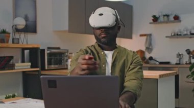 Diz üstü bilgisayara bağlı vr kulaklık takan Afrikalı Amerikalı iş adamının evde gelişmiş gerçeklikte çalışırken çekilmiş orta boy fotoğrafı.