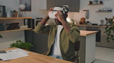 Siyah girişimcinin sanal gerçeklik kulaklığı taktığı ve evden çevrimiçi çalışırken el hareketleriyle hareket ettiği bir fotoğraf.