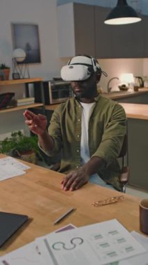 Afro-Amerikalı iş adamının evdeki masada el kol hareketleri kullanarak sanal gerçeklik gözlükleri içinde çalışmasının dikey çekimi.