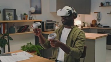 VR kulaklık takan ve evdeki masadaki artırılmış gerçeklikte çalışırken kontrol cihazları kullanan Afrikalı Amerikalı girişimcinin arkası.