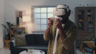 Genç adamın sanal gerçeklik gözlüğü ve kumandayla rakiplerine saldırdığı orta boy bir fotoğraf. Oturma odasının ortasında duruyor.