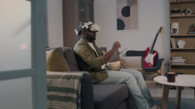 Arttırılmış gerçeklik kulaklığı takmış neşeli siyahi adamın film seçerken ve evdeki kanepede patlamış mısır yerken yan pozunu çek.