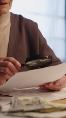 Yaşlı kadın emeklinin gecikmiş ödemeler, faturalar ve masadaki nakitle uğraşırken kamu hizmeti sağlayıcısı sözleşmesinde yüksek sesle küçük yazılar okuduğu orta boy dikey bir orta kesim çekimi.