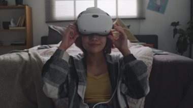 Kızıl saçlı Asyalı genç kızın orta boy portresi. Sanal gerçeklik gözlüklerini çıkarıyor ve evde yatağın yanında poz veriyor, kameraya bakıyor.