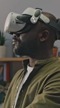 Siyah adamın VR gözlüklü dikey görüntüsü artırılmış gerçekliğe daldırılmış, evde çalışıyor.