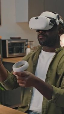 VR kulaklık takan ve evdeki masadaki artırılmış gerçeklikte çalışırken kontrolör kullanan Afrikalı Amerikalı girişimcinin dikey yay görüntüsü.