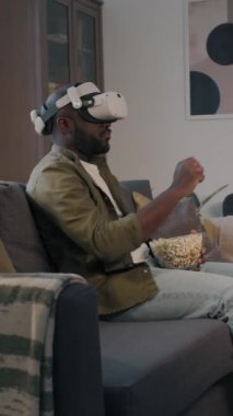 Arttırılmış gerçeklik kulaklığı takmış neşeli siyahi adamın evde kanepede patlamış mısır yiyip film seçerken dikey yan görüntüsü.