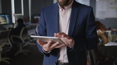 Kendine güvenen inek Arap erkek STEM hocasının tablet kullanan ve kameraya bakan orta ölçekli eğik portresi ve bilgisayar başında oturmuş kodlama ödevi üzerinde çalışan genç öğrenciler.